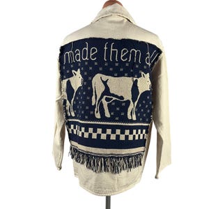 May include: A cream-colored long-sleeve shirt featuring a large, rectangular navy blue woven panel on the back. The panel displays two cows, a checkered border, fringe at the bottom, and the text "made them all" in white cursive lettering.