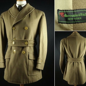 Puede incluir: Un abrigo de lana marrón de doble botonadura con cinturón, bolsillos grandes y cuello ancho. El abrigo tiene botones dorados y una etiqueta que dice "Abercrombie & Fitch Co. NEW YORK".