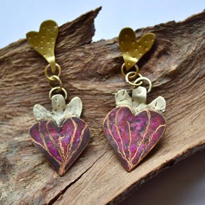 Puede incluir: Pendientes hechos a mano con colgantes en forma de corazón en morado y dorado. Los pendientes tienen una parte superior dorada en forma de corazón y están conectados por pequeños anillos dorados. El diseño es intrincado.