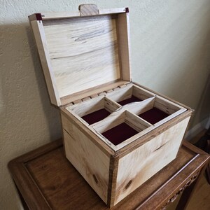 May include: A wooden box with a hinged lid, open to reveal four interior compartments lined with red fabric. The box is made of light-colored wood with darker wood trim and a wooden latch. The box is sitting on a wooden table.