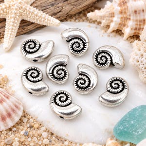 May include: A collection of eight silver-toned metal beads shaped like nautilus shells. Each bead features a detailed spiral pattern with dark recessed grooves and a smooth, polished outer surface. The beads are arranged in a cluster on a white surface.