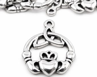 May include: A silver-toned metal Claddagh charm featuring a heart topped with a crown, held by two hands, and surmounted by a triquetra knot. The charm includes a small circular loop at the top for attachment, with additional charms blurred in the background.