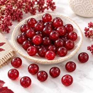 May include: A collection of round, translucent red beads with internal glitter particles. Many beads are clustered inside a small, speckled, off-white ceramic bowl, while several individual beads are scattered on the surface around the bowl.