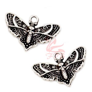 Puede incluir: Dos colgantes de mariposa plateados con intrincados diseños de alas y un lazo en la parte superior para colgar. Los colgantes tienen un aspecto detallado y texturizado, con un acabado oscuro y antiguo. Las alas de mariposa presentan un patrón de puntos y líneas curvas.