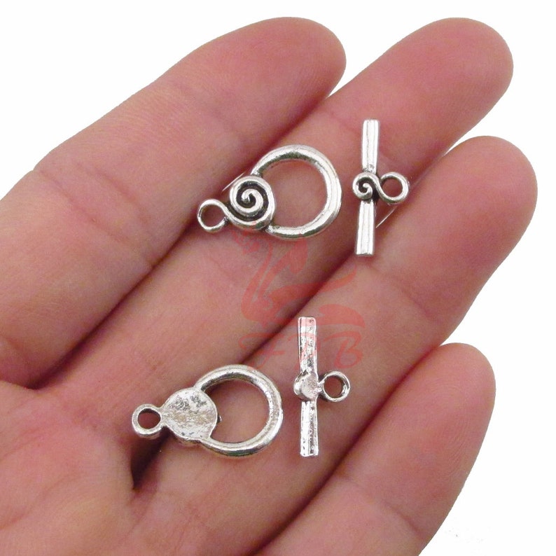 Puede incluir: Dos cierres de palanca plateados con un dise&ntilde;o decorativo de espiral. Cada cierre tiene un bucle redondo y una barra recta con un peque&ntilde;o bucle en el extremo.