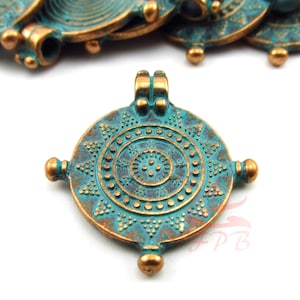 May include: A round, teal-colored metal pendant with a decorative design of concentric circles and triangles. The pendant has a loop at the top for attaching to a chain or cord.