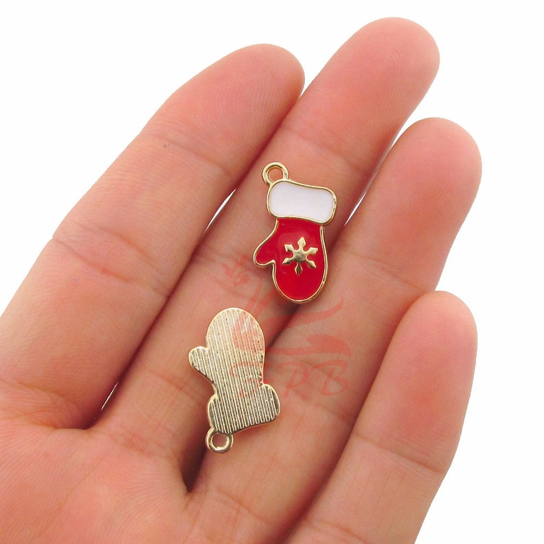 May include: Two gold-tone metal charms shaped like mittens. One mitten is red with white trim and a snowflake design. The other mitten is gold with a textured surface.