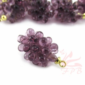 May include: A cluster of purple glass beads shaped like grapes. The beads are a translucent purple color, with a gold-colored metal loop attached. The grapes are on a white background.