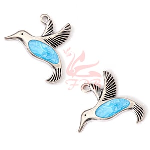Op de afbeelding: Twee zilverkleurige kolibri-bedels met blauwe emaille details. De bedels hebben gedetailleerde vleugels en een lus om aan sieraden te bevestigen. Het blauwe emaille heeft een gemarmerd patroon. De bedels staan tegen een witte achtergrond.