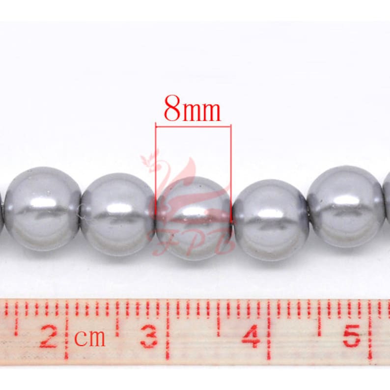 May include: A strand of 8mm silver-grey faux pearl beads. The beads are round and smooth, and they are strung on a clear thread. The beads are measured against a ruler, showing that they are 8mm in diameter.