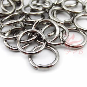 May include: A collection of shiny, silver-colored jump rings. The rings are round with a small opening, and are scattered across a white surface. The jump rings are made of metal and reflect light.