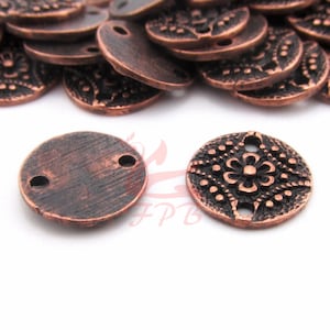 Op de afbeelding: Een stapel ronde metalen bedels in koperkleur met twee gaten. Sommige bedels hebben een bloemmotief en andere zijn effen.