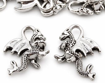 May include: A collection of silver-toned metal dragon charms, each featuring a detailed scaled body, outstretched wings, and a small loop at the top for attachment. The charms are arranged with two in the foreground and several others blurred in the back.