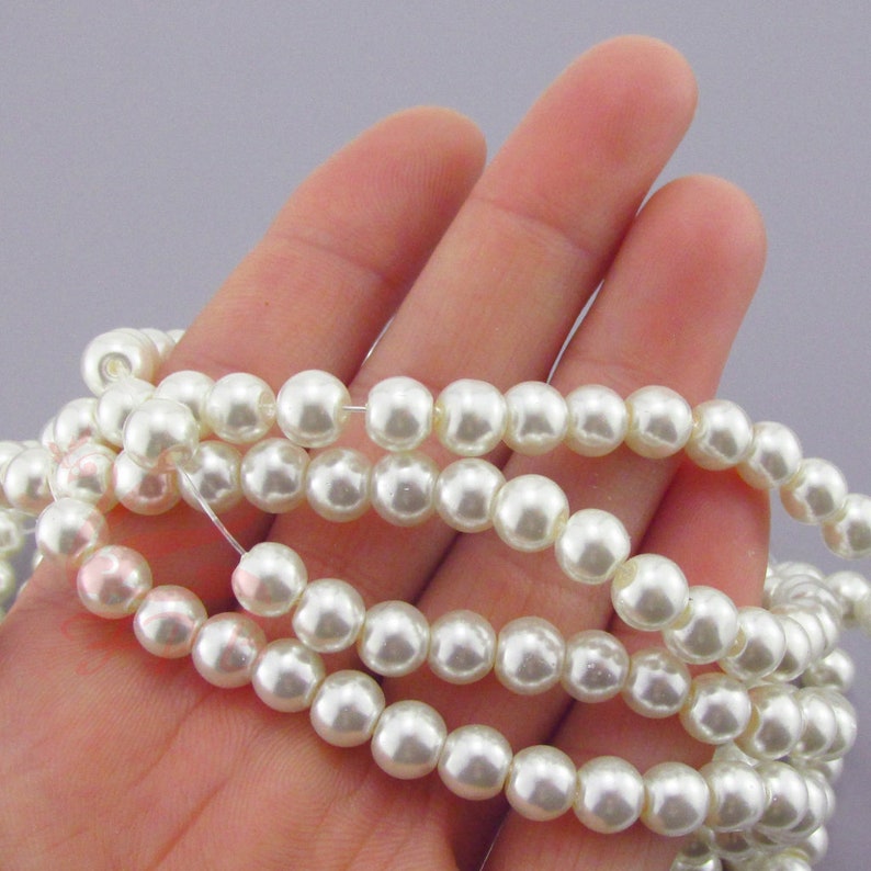 May include: A strand of white pearl beads, shown in a hand, for jewelry making.
