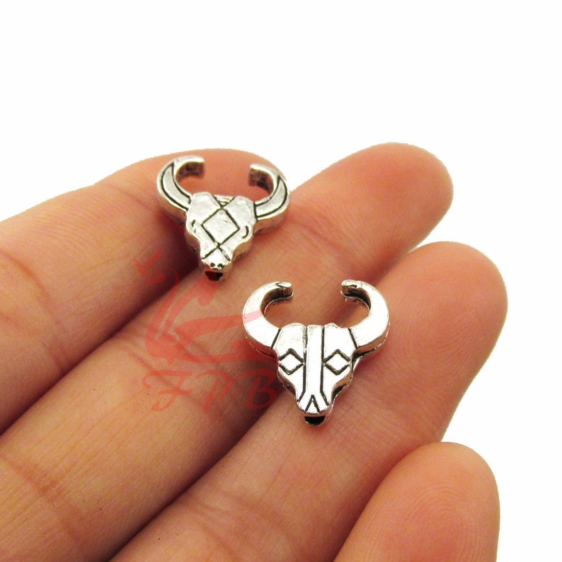May include: Two silver bull head charms with geometric designs. The charms are on a hand.