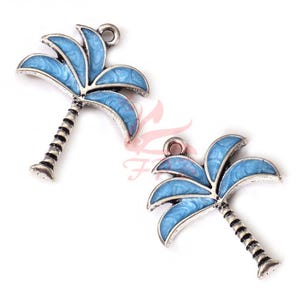 Op de afbeelding: Twee zilverkleurige palmboombedels met blauwe emaille details. De palmbladeren zijn lichtblauw en de stammen zwart en zilver. Elke bedel heeft een kleine lus aan de bovenkant om te bevestigen.