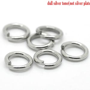 May include: Six silver jump rings, also known as split rings, are shown on a white surface. The rings are made of metal and have a dull silver tone. The rings are open and ready to be used for jewelry making or other crafts.