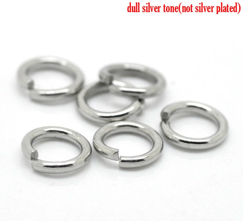 May include: Six silver jump rings, also known as split rings, are shown on a white surface. The rings are made of metal and have a dull silver tone. The rings are open and ready to be used for jewelry making or other crafts.
