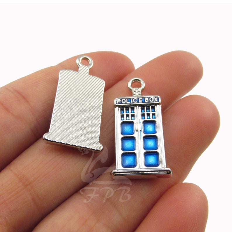 K&ouml;nnte beinhalten: Silberner Anh&auml;nger mit einem blauen und wei&szlig;en Emaille-Polizeikasten-Design. Der Anh&auml;nger tr&auml;gt die Aufschrift "Police Box" auf der Vorderseite. Die R&uuml;ckseite des Anh&auml;ngers ist schlicht silber.