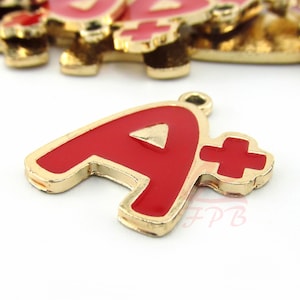 May include: Gold-colored metal charm with a red enamel letter A and a plus sign. The charm is shaped like a letter A with a red enamel fill and a gold outline. The plus sign is also red enamel with a gold outline.