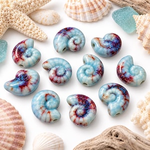 May include: A collection of ten ceramic beads shaped like nautilus shells, featuring a glossy light blue glaze with dark red and purple crackle patterns. Each bead has a central hole for stringing, arranged among various natural seashells.