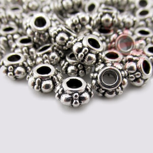 May include: Close-up of a pile of silver-colored metal beads. Each bead has a flower-like design with a large central hole and smaller decorative bumps around the edges. The beads are scattered on a white surface.