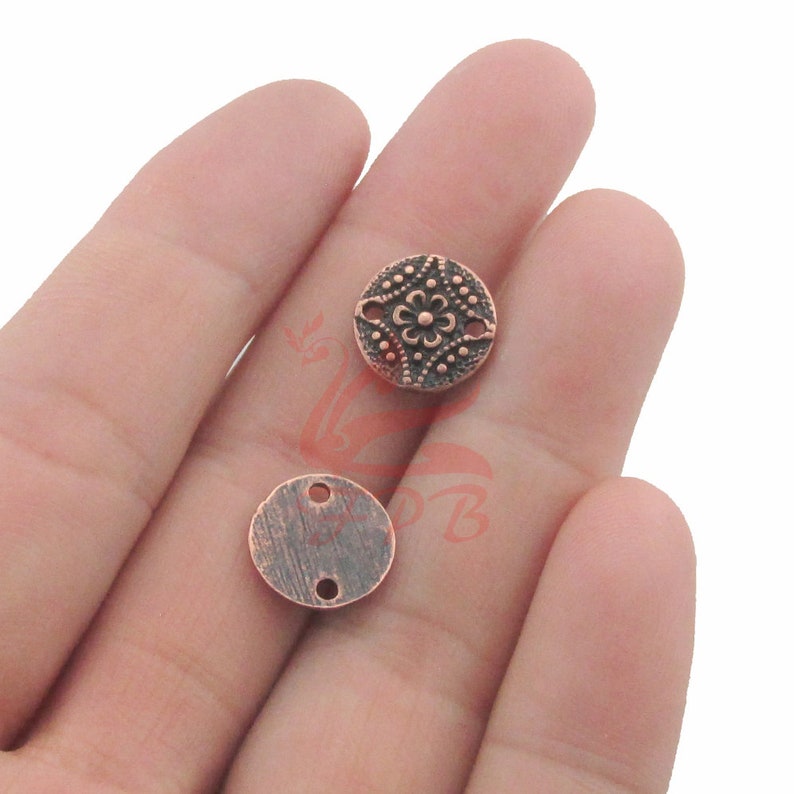 Op de afbeelding: Twee koperkleurige ronde metalen kralen met een decoratief ontwerp. E&eacute;n kraal heeft een bloemmotief en de andere is effen. Beide kralen hebben twee gaten om te rijgen.