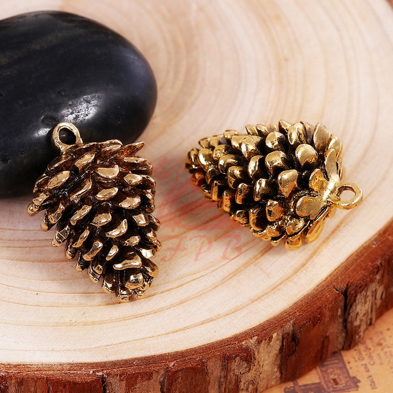 May include: Two gold-toned metal pine cone charms with a loop for hanging. The pine cones are detailed and realistic.