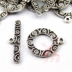 May include: Silver-toned metal toggle clasp with a decorative swirl pattern. The clasp has a round ring and a straight bar with a loop at one end.