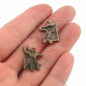 May include: Two antique gold-toned metal charms featuring Egyptian-inspired designs. One charm depicts an anubis jackal head with a detailed face and a textured mane. The other charm depicts a stylized anubis jackal standing on four legs.