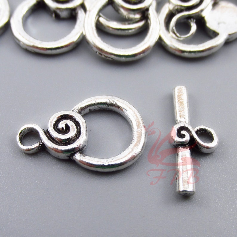 Puede incluir: Cierre de metal plateado con un dise&ntilde;o de espiral decorativo. El cierre tiene un anillo redondo y una barra recta con una espiral en un extremo.