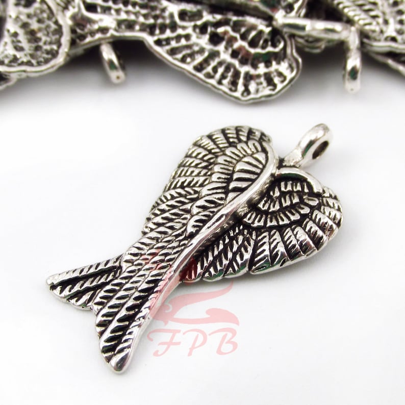 May include: Silver charm featuring a detailed design of angel wings in a heart shape. The charm has a loop at the top for attaching to a necklace or bracelet.