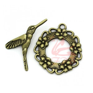 May include: A set of two antique gold-toned metal charms. One charm is a hummingbird with detailed wings and a long tail. The other charm is a circular frame with a floral design and a cutout center.