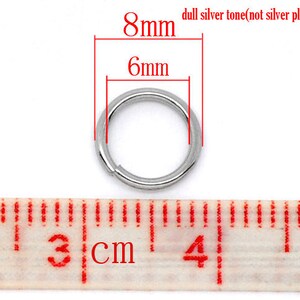 May include: A silver-toned metal jump ring measuring 8mm in diameter and 6mm in width. The ring is shown next to a ruler, with the measurement markings visible.