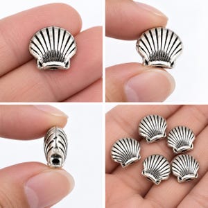 May include: A collection of silver-toned metal beads shaped like seashells. Each bead features a detailed ribbed texture with dark oxidized grooves. The beads are small, rounded, and include a visible hole at the base for threading onto jewelry.