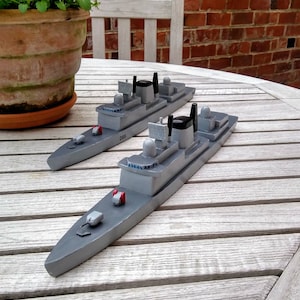 May include: Two grey wooden toy battleships with black and white details sit on a weathered wooden table. The ships have small red and white accents and are labelled with the number "D89". A plant in a terracotta pot and a white chair are in the background.