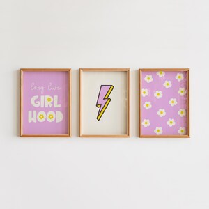 Op de afbeelding: Drie rechthoekige wandkunstprints in licht houten lijsten. De linker print heeft een paarse achtergrond met de witte tekst "long live GIRL HOOD" en een klein geel bloemetje. De middelste print toont een paarse bliksemschicht met een gele omlijning op een crèmekleurige achtergrond. De rechter print toont een herhalend patroon van witte bloemen met gele harten op een paarse achtergrond.
