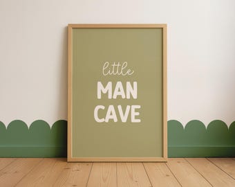 May include: A rectangular wall art print in a light wood frame featuring a muted olive green background with the text "little MAN CAVE" printed in white. The word "little" is in a thin script font, while "MAN CAVE" is in a bold, sans-serif font.