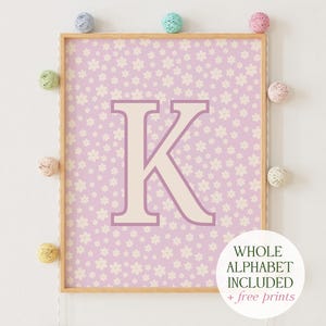 May include: A framed print with a light purple background and a white letter K in the center. The background is covered in a pattern of small white flowers. The text "WHOLE ALPHABET INCLUDED + free prints" is at the bottom of the print.