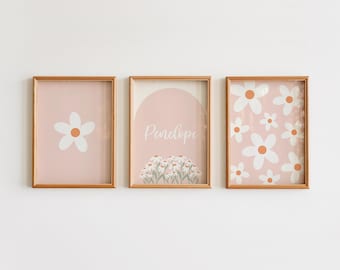 Puede incluir: Tres cuadros rectangulares con marcos de madera clara. El de la izquierda muestra una flor blanca sobre fondo rosa empolvado. El central presenta el nombre Penelope sobre un grupo de margaritas. El de la derecha tiene un patrón de flores.