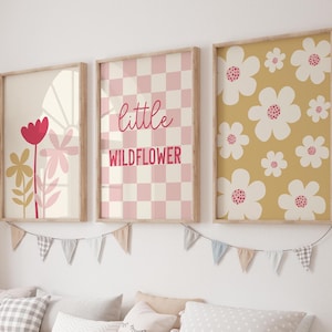 May include: Three framed prints with a floral theme. The left print features a pink and yellow flower with a red center. The middle print has a pink and white checkered background with the text "little wildflower". The right print has a yellow background with white flowers with pink centers.