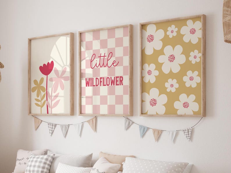 May include: Three framed prints with a floral theme. The left print features a pink and yellow flower with a red center. The middle print has a pink and white checkered background with the text "little wildflower". The right print has a yellow background with white flowers with pink centers.