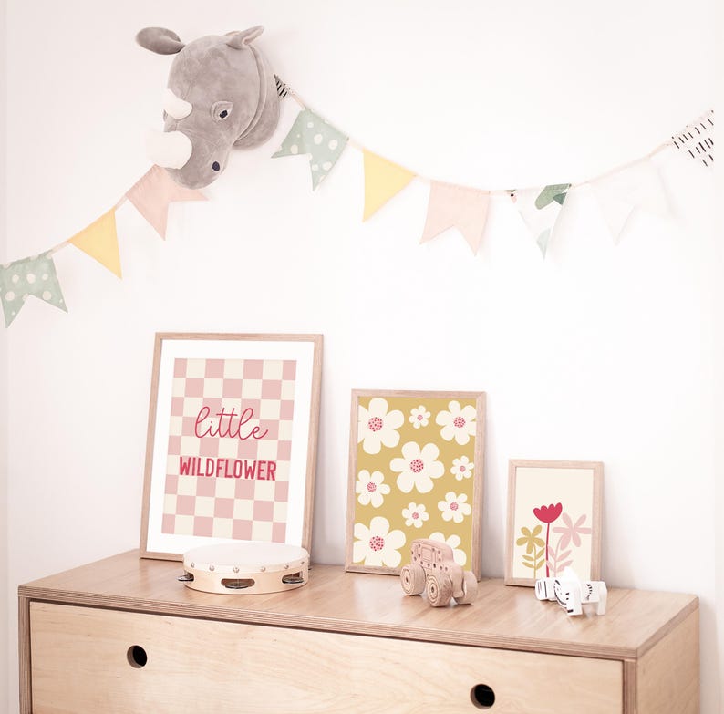 May include: A nursery room decor set with a gray stuffed rhino head, a pink and white checkered print with the text "little wildflower", a yellow and white floral print, and a red and green floral print. The prints are framed and hung on a white wall. A wooden toy truck and a tambourine are on a wooden dresser.