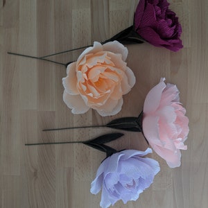 May include: Four crepe paper peony flowers in shades of peach, pink, purple, and burgundy. Each flower has a green wire stem and dark green leaves. The flowers are arranged on a light wood grain surface.