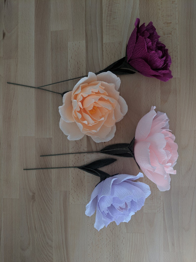 May include: Four crepe paper peony flowers in shades of peach, pink, purple, and burgundy. Each flower has a green wire stem and dark green leaves. The flowers are arranged on a light wood grain surface.