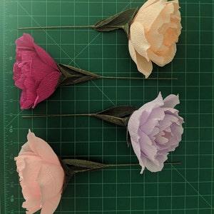 May include: Four crepe paper peony flowers in shades of pink, peach, and lavender, with green stems and leaves. The flowers are arranged on a green cutting mat, showcasing their delicate, textured petals.