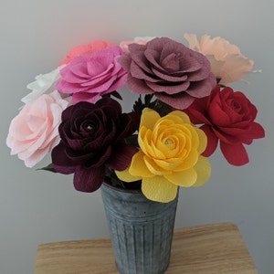 May include: A bouquet of colorful paper flowers in a galvanized metal bucket. The flowers are various shades of pink, red, yellow, and purple. The bucket sits on a wooden surface.