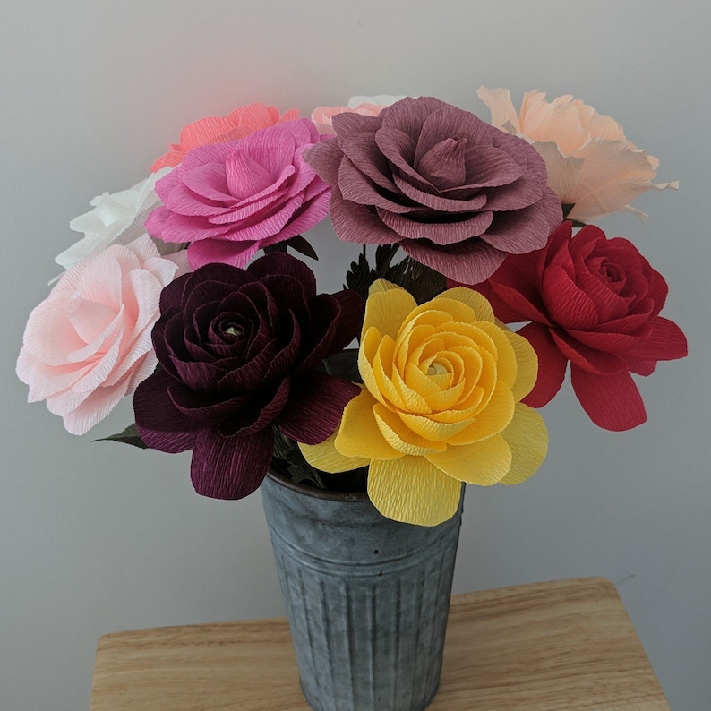 May include: A bouquet of colorful paper flowers in a galvanized metal bucket. The flowers are various shades of pink, red, yellow, and purple. The bucket sits on a wooden surface.
