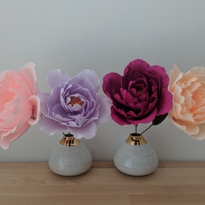 May include: Four large paper flowers in shades of pink, purple, and burgundy, displayed in small white vases with gold rims. The flowers are arranged on a light wooden surface against a neutral background.