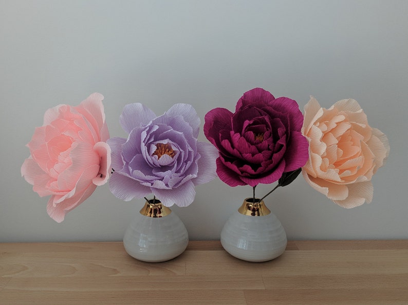 May include: Four large paper flowers in shades of pink, purple, and burgundy, displayed in small white vases with gold rims. The flowers are arranged on a light wooden surface against a neutral background.